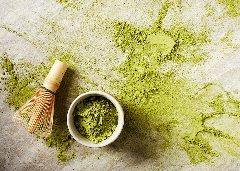 Navitas Organics Matcha Powder in a dish next to a wooden whisk.