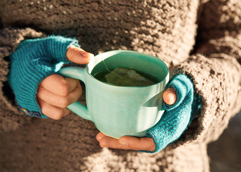 A woman in a heavy coat and gloves holding a mug of warm tea.