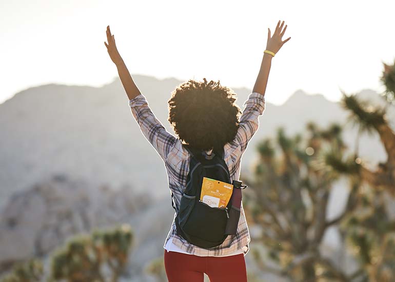 A woman with a backpack full of Navitas Organics Goldenberries raising her arms as she faces the sun in the desert.
