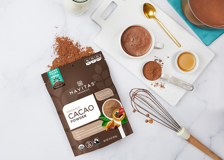 Navitas Organics Cacao Powder with a whisk, plant-based milk and honey on a cutting board