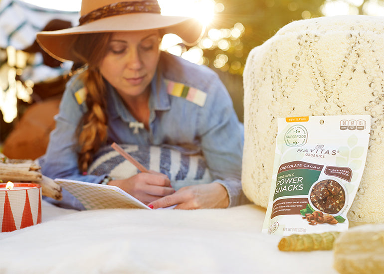 A woman writing her New Year's resolutions in a journal on a campsite with a bag of Navitas Organics Chocolate Cacao Power Snacks.