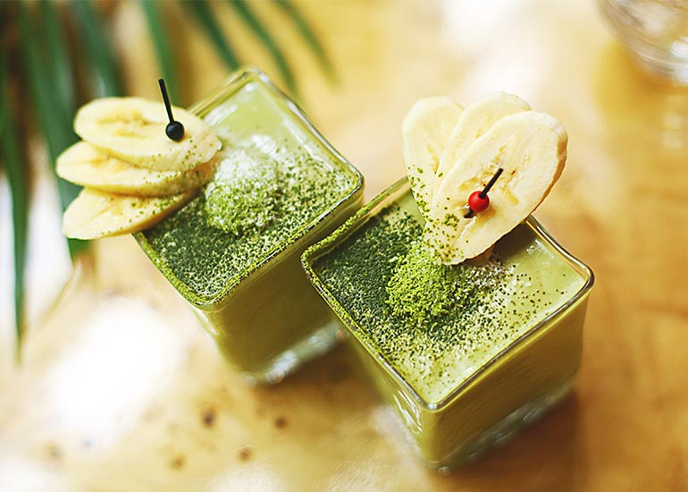 Two tall serving glasses filled with creamy blended matcha smoothies made with Navitas Organics Matcha Powder, topped with fresh banana slices.