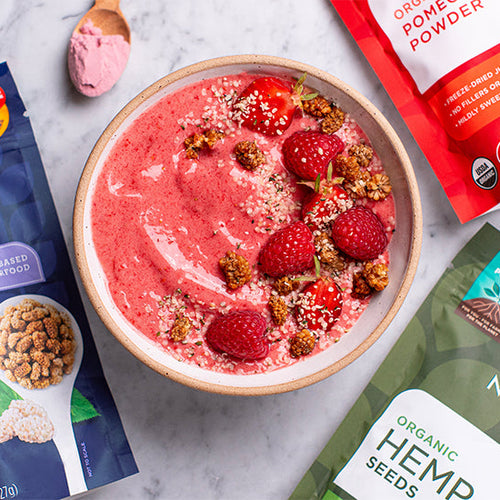 A pink smoothie bowl topped with raspberries, hemp seeds and mulberries, surrounded by various Navitas Organics products including hemp seeds and goji berries.