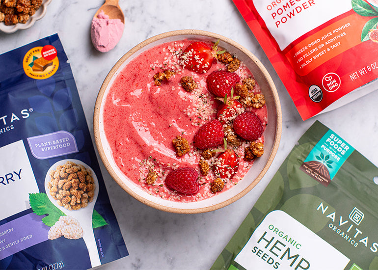 A pink smoothie bowl topped with raspberries, hemp seeds and mulberries, surrounded by various Navitas Organics products including hemp seeds and goji berries.