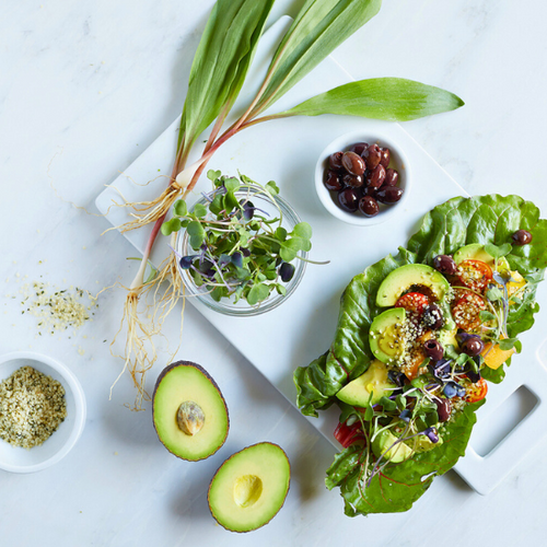 A healthy meal setup including a plate of leafy greens topped with sliced avocado, microgreens, and seeds, accompanied by bowls of sprouts, olives, and hemp seeds on a marble surface.