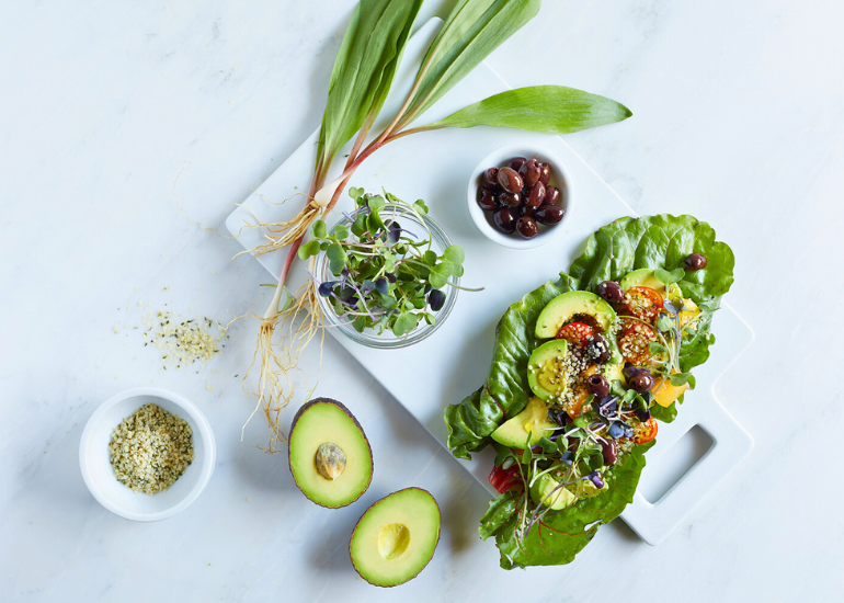 A healthy meal setup including a plate of leafy greens topped with sliced avocado, microgreens, and seeds, accompanied by bowls of sprouts, olives, and hemp seeds on a marble surface.