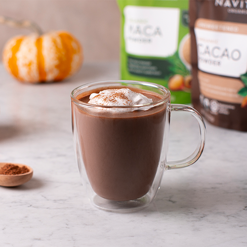 A cup of hot chocolate topped with whipped cream and a sprinkle of cacao powder, with bags of Navitas Organics Cacao Powder and Maca Powder visible in the background, next to a small pumpkin and a wooden spoon filled with cacao powder.