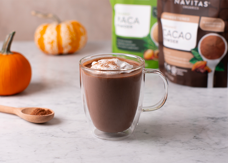 A cup of hot chocolate topped with whipped cream and a sprinkle of cacao powder, with bags of Navitas Organics Cacao Powder and Maca Powder visible in the background, next to a small pumpkin and a wooden spoon filled with cacao powder.