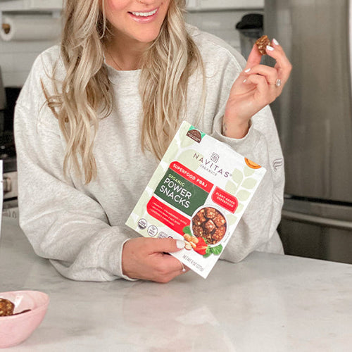 A person holding a bag of Navitas Organics Superfood PB&J Power Snacks, smiling while sitting at a kitchen counter with a bowl of snacks and another product bag visible.
