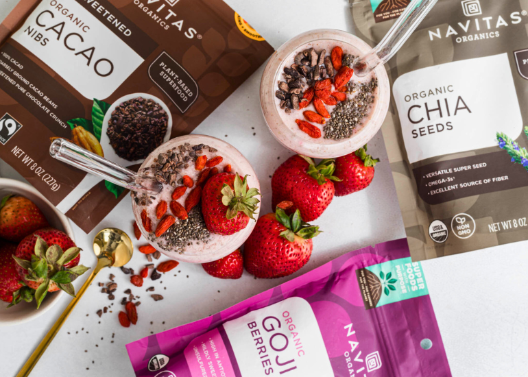 Two smoothie glasses topped with chia seeds, cacao nibs, and goji berries are surrounded by fresh strawberries. Nearby are packages of Navitas Organics products, including Cacao Nibs, Chia Seeds, and Goji Berries. A golden spoon is placed on the side.