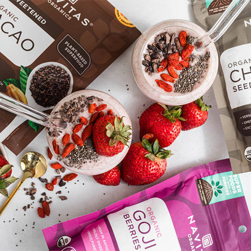 Two smoothie garnished with strawberries, cacao nibs, and chia seeds, accompanied by spoons and surrounded by bags of Navitas Organics Cacao Nibs, Chia Seeds and Goji Berries.