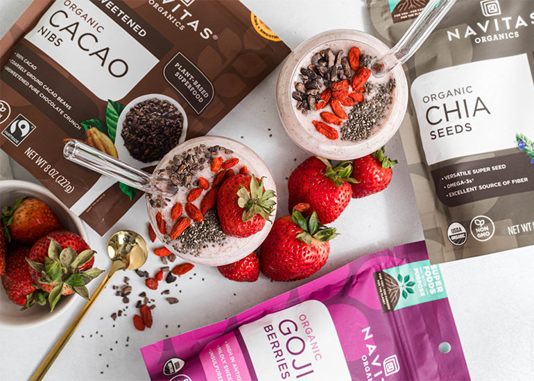 Two smoothie garnished with strawberries, cacao nibs, and chia seeds, accompanied by spoons and surrounded by bags of Navitas Organics Cacao Nibs, Chia Seeds and Goji Berries.