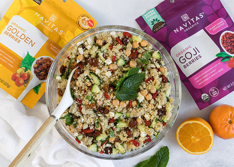 A bowl of quinoa salad with mint leaves, beans and dried fruits next to bags of Navitas Organics Goldenberries and Goji Berries, with sliced oranges on the side.