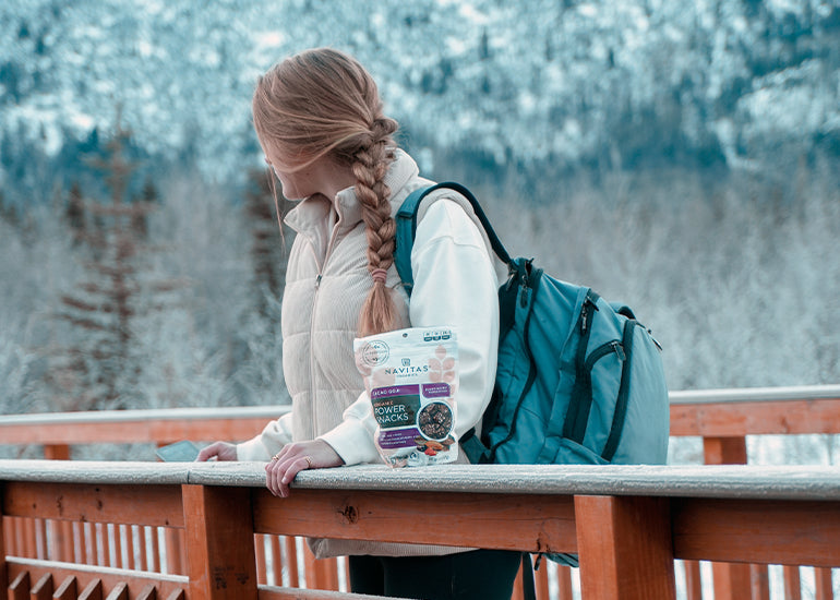 A woman with a backpack standing on a bridge behind a bag of Navitas Organics Cacao Goji Power Snacks