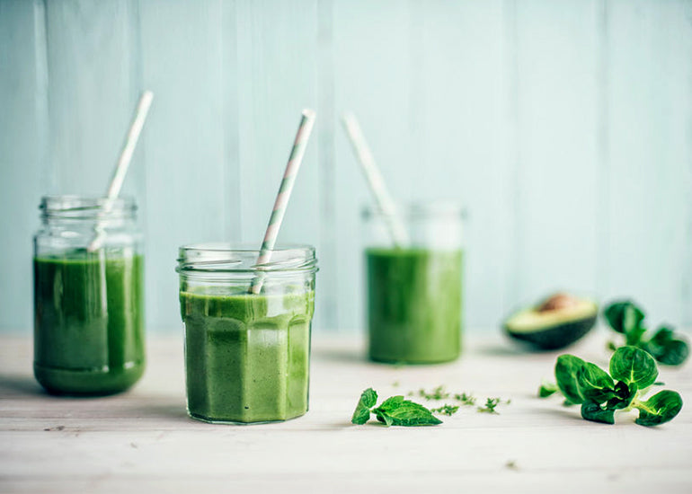 Three green smoothies in glasses with various plants, herbs and an avocado surrounding them