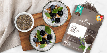 Two dishes filled with chia pudding topped with berries, nuts and mint leaves next to a dish filled with chia seeds and a spilling bag of Navitas Organics Chia Seeds.