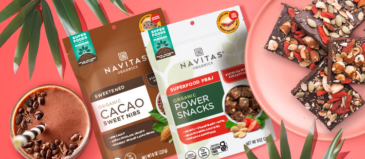 Bags of Navitas Organics Cacao Sweet Nibs and PB&J Power Snacks beside a chocolate smoothie, a plate topped with nut and dried fruit covered chocolate bark and palm leaves against a coral colored background. 