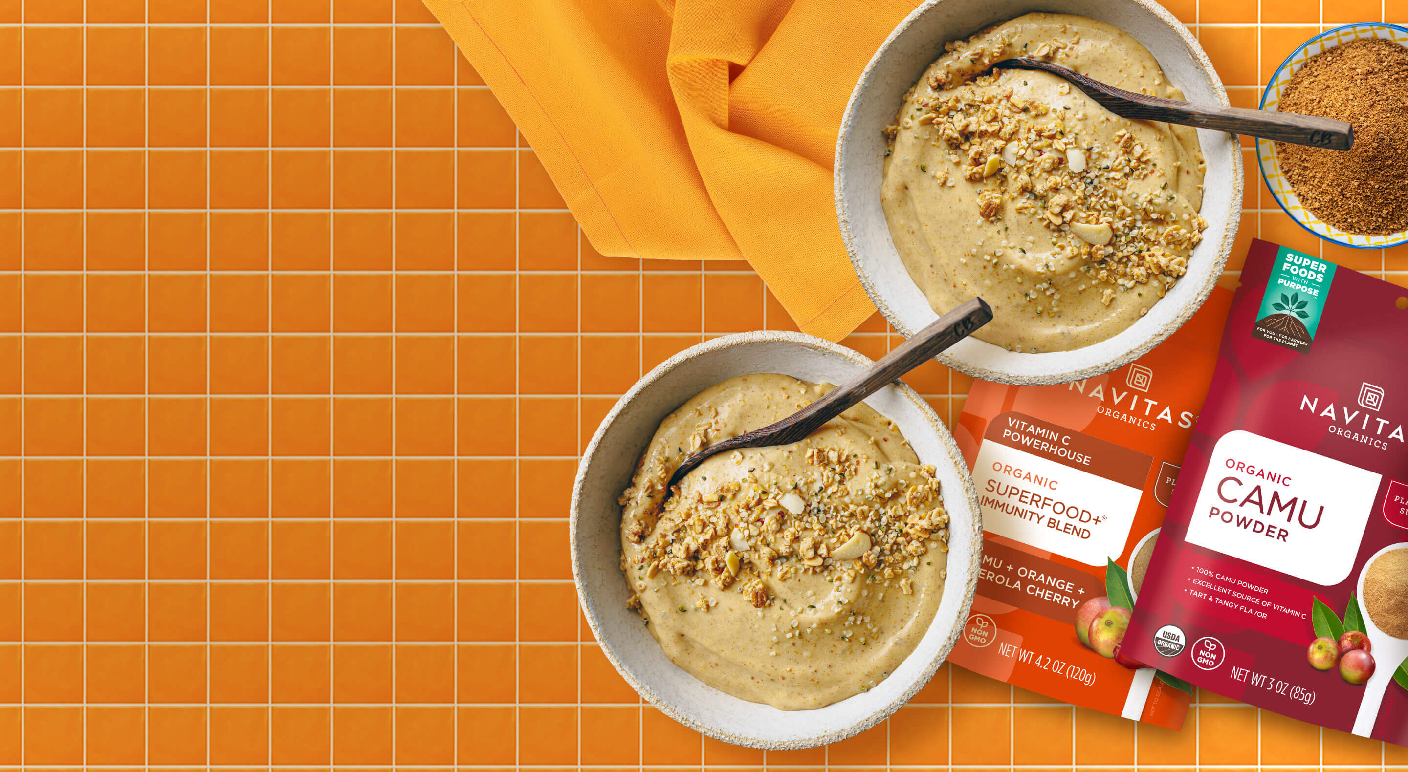 An orange countertop with two smoothie bowls, a dish filled with organic camu powder and bags of Navitas Organics Superfood+ Immunity Blend and Camu Powder.