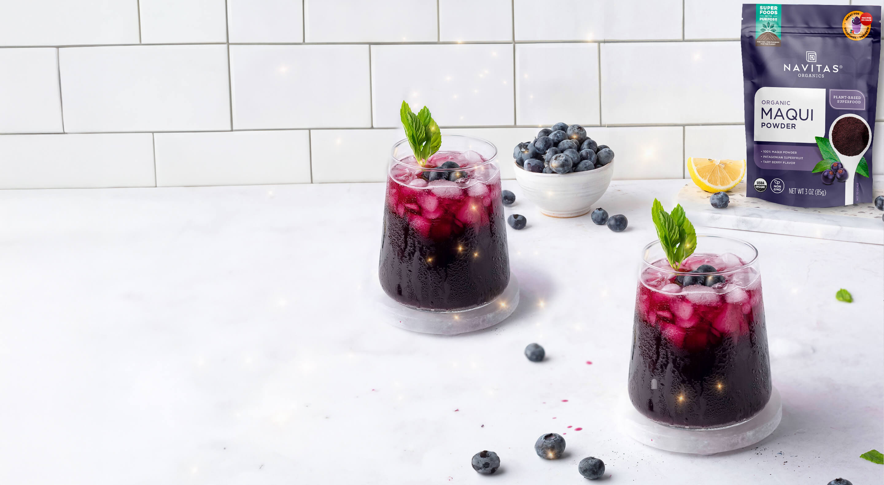 Two glasses filled with vibrant purple drinks garnished with fresh mint and blueberries with a small bowl of blueberries and a bag of Navitas Organics Maqui Powder visible in the background.