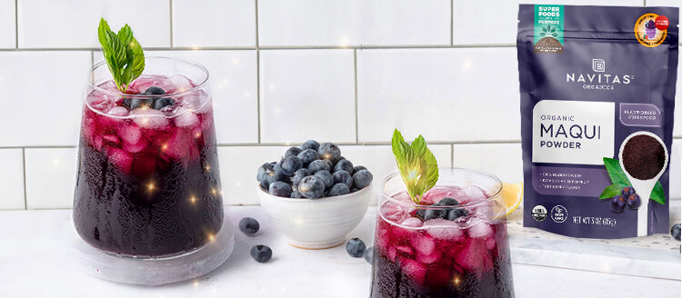 Two glasses filled with vibrant purple drinks garnished with fresh mint and blueberries with a small bowl of blueberries and a bag of Navitas Organics Maqui Powder visible in the background.