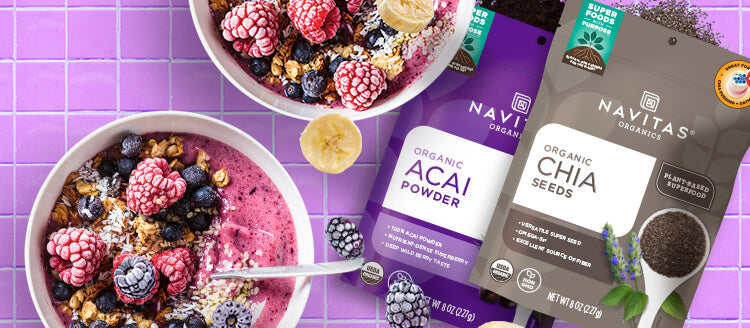 Two smoothie bowls topped with various berries, coconut and granola next to bags of Navitas Organics Acai Powder and Chia Seeds bags on a pink tile countertop.