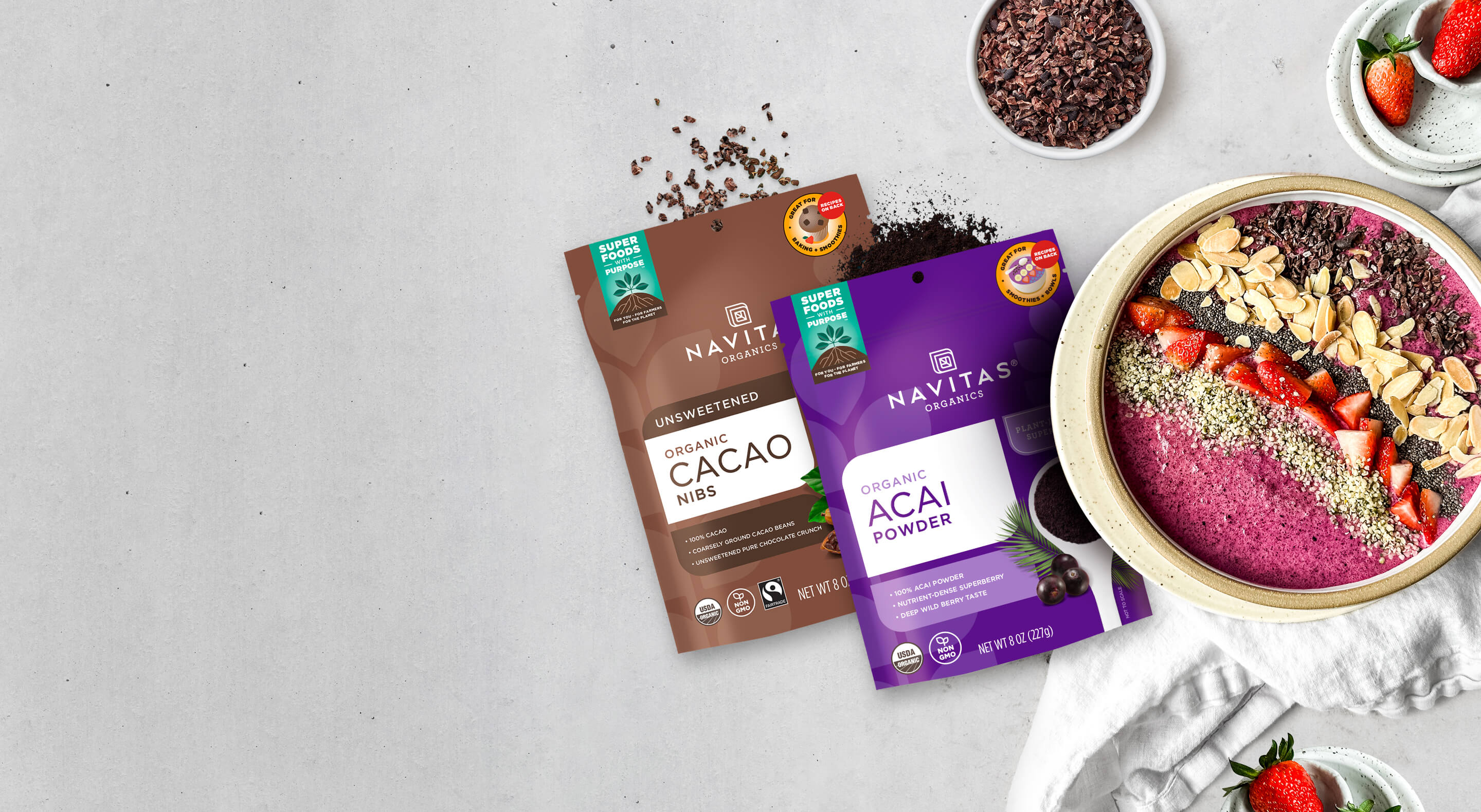 An overhead shot of a smoothie bowl topped with various nuts, seeds, berries and cacao nibs surrounded by bags of Navitas Organics Cacao Nibs and Acai Powder, a dish filled with cacao nibs and dishes topped with fresh strawberries.