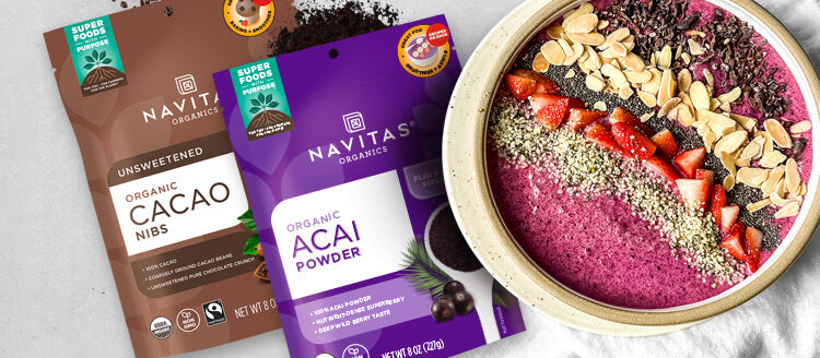 An overhead shot of a smoothie bowl topped with various nuts, seeds, berries and cacao nibs surrounded by bags of Navitas Organics Cacao Nibs and Acai Powder.
