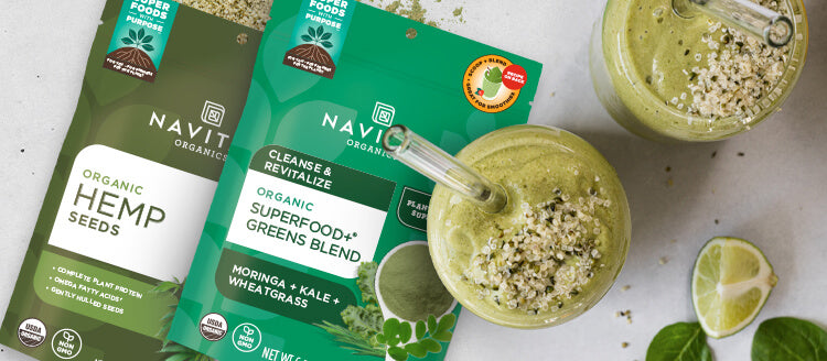 Two green smoothies topped with hemp seeds next to bags of Navitas Organics Hemp Seeds and Superfood+ Greens Blend, and a fresh lime.