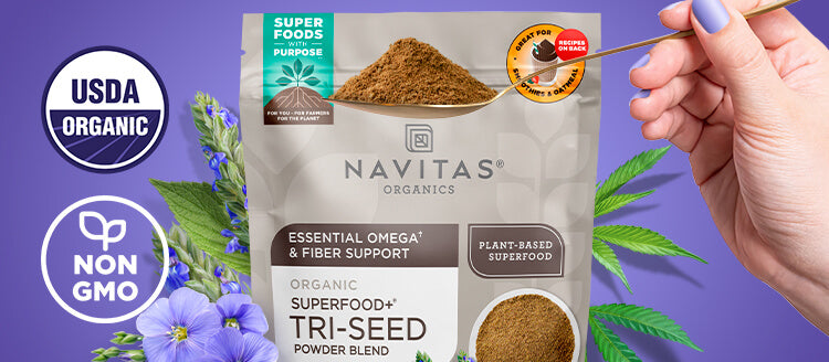 Navitas Organics Superfood+ Tri-Seed Powder Blend package with a hand holding a spoonful of powder against a purple background.