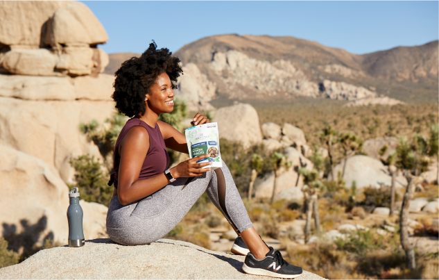 Young lady sitting on a rock overlooking a dessert backdrop with a drinking bottle and package of Navitas Superfood snacks.