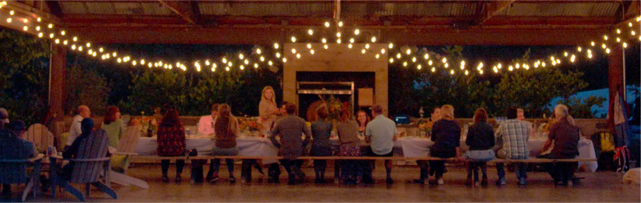 Long table with strings of lights overhead with people eating together.